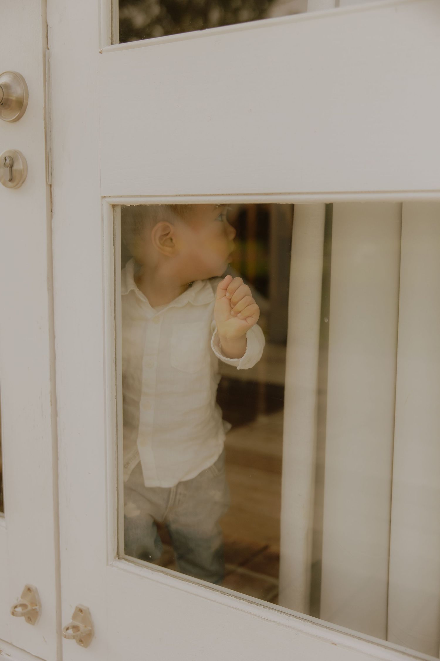 Curious toddler in white linen shirt peers through glass panels of a white door.