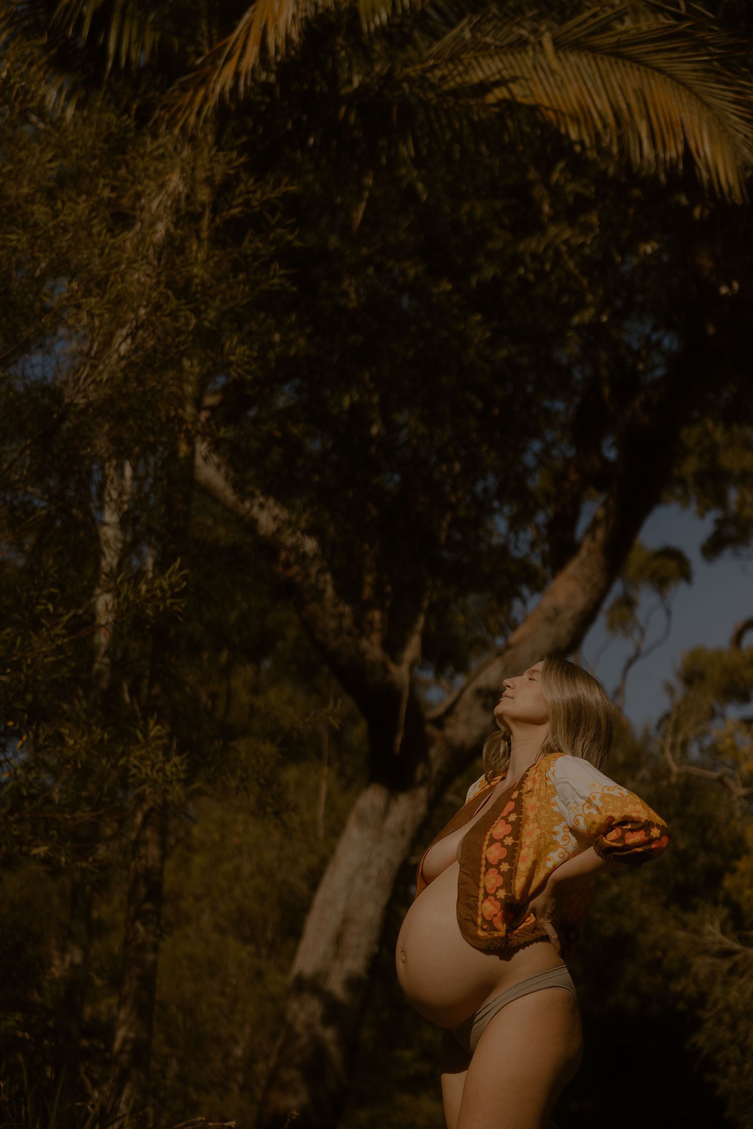 Pregnant woman in swimwear tilting her head back among lush tropical trees in golden light.