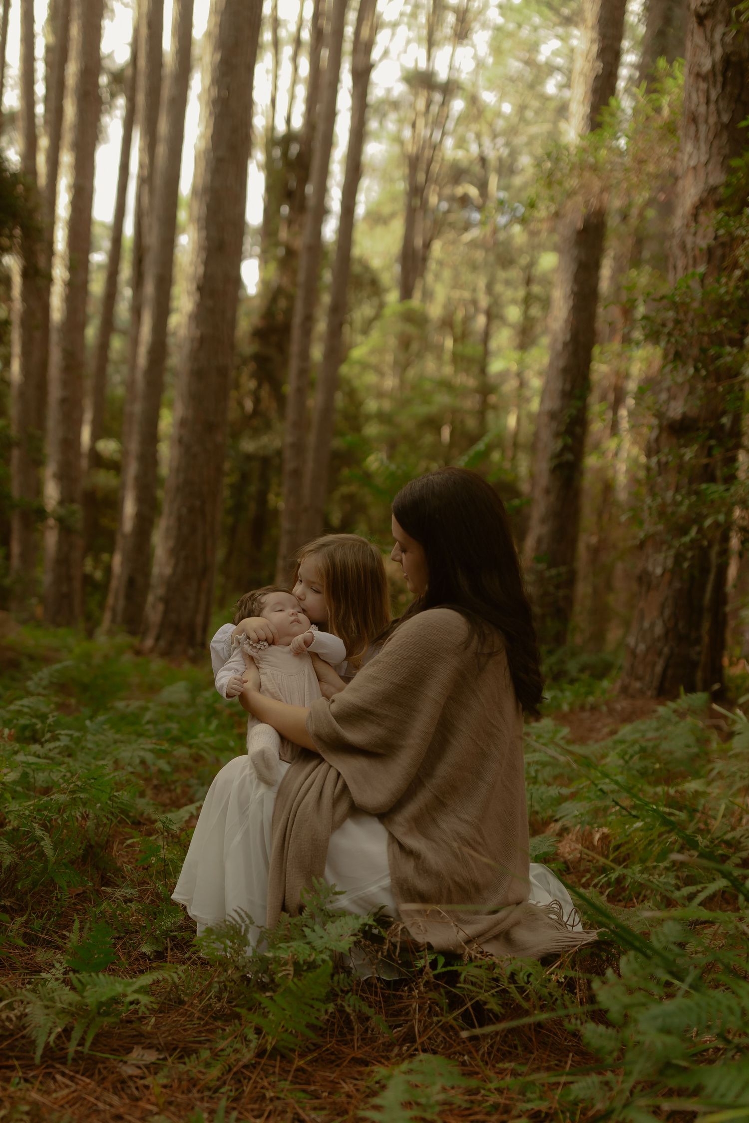 Mother tenderly embraces young daughter while sitting among green ferns in a forest.