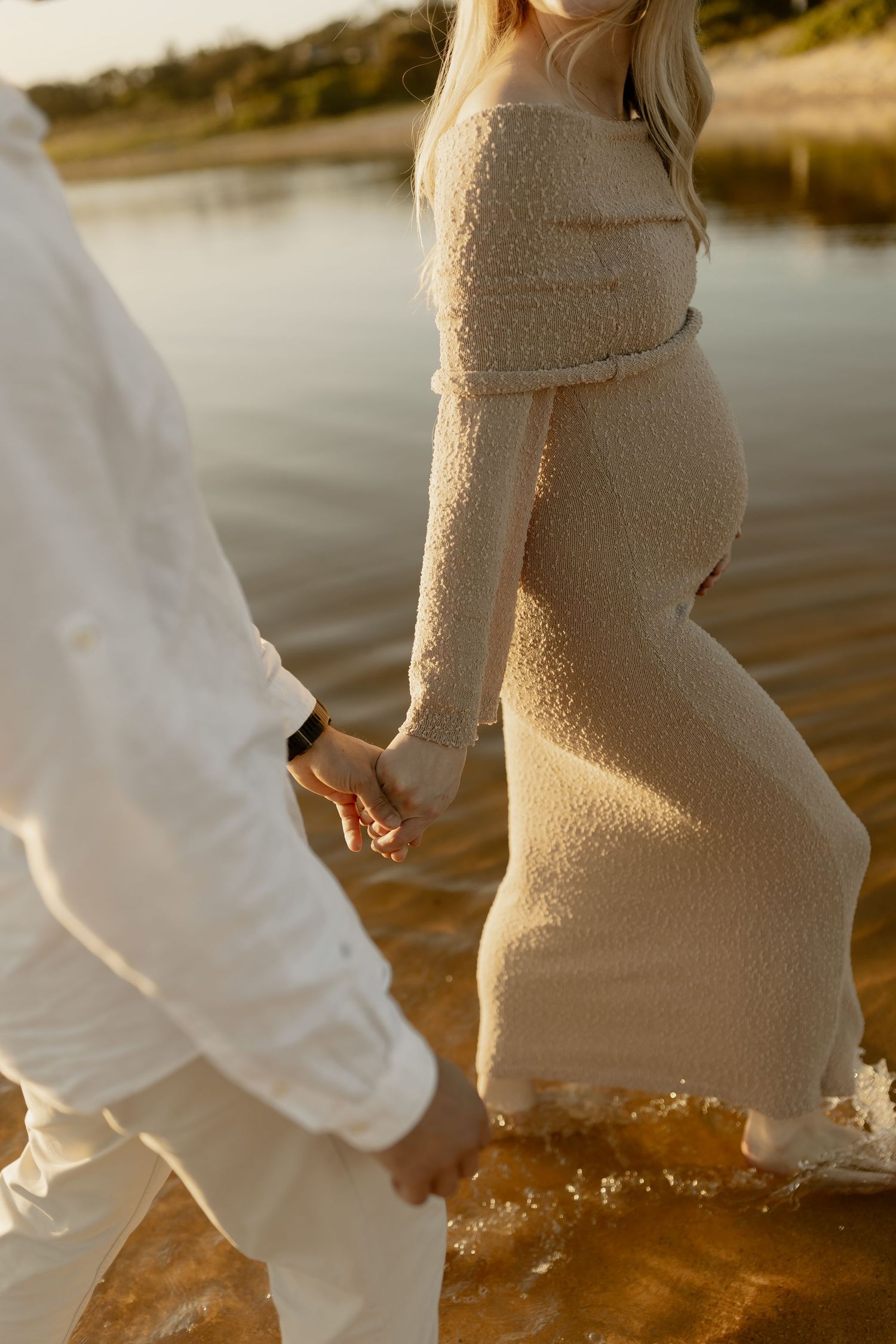 Couple holding hands while standing in shallow water at sunset wearing cream colored outfits.