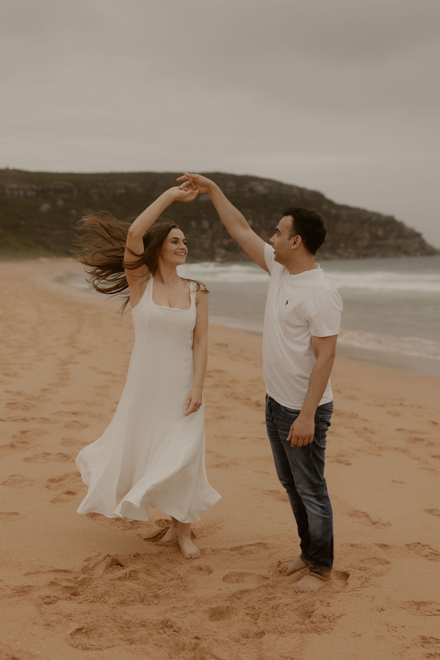 A couple in white and casual attire dances joyfully together on a beach with cliffs in the distance.