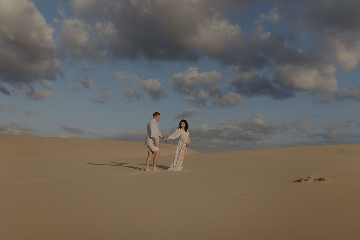 A couple holds hands while walking through a vast desert landscape under dramatic cloudy skies at sunset.