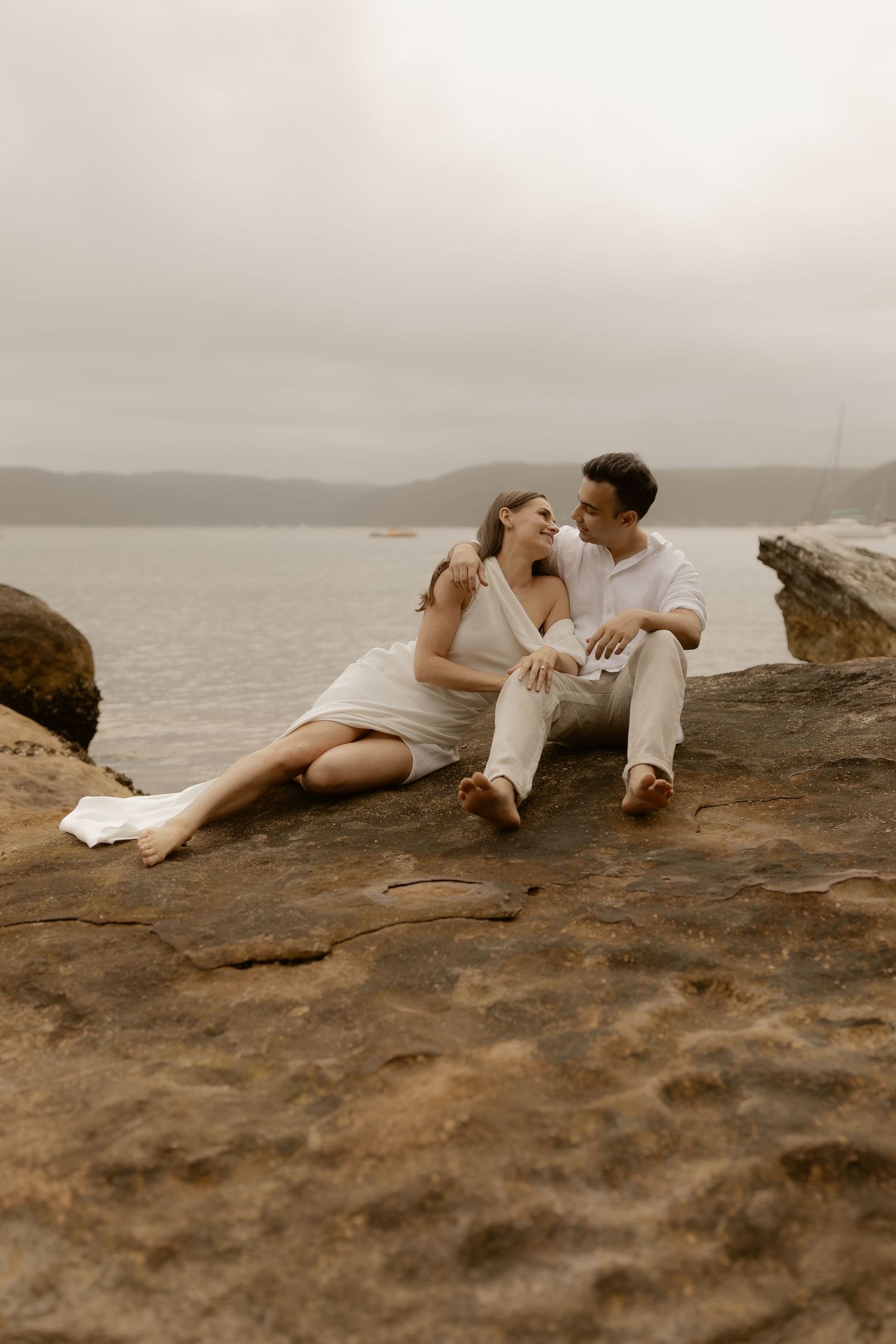 A romantic couple in white clothing lounges on coastal rocks overlooking the ocean on an overcast day.