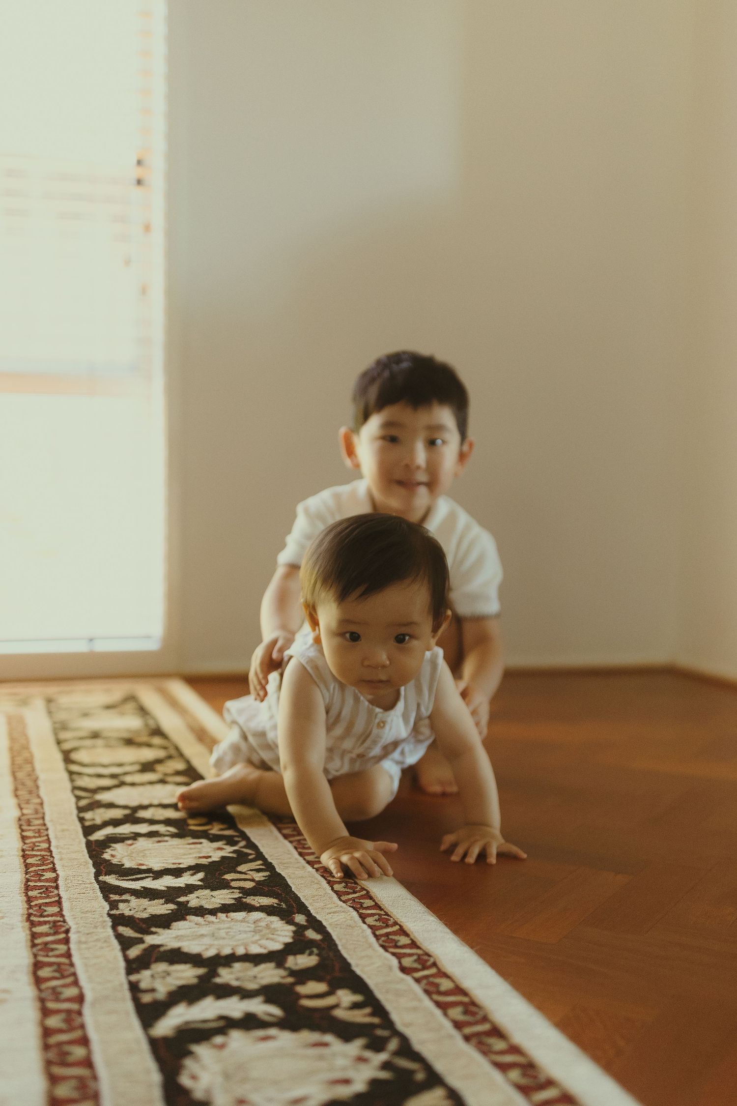 Two children play together on a patterned rug in a sunlit room.