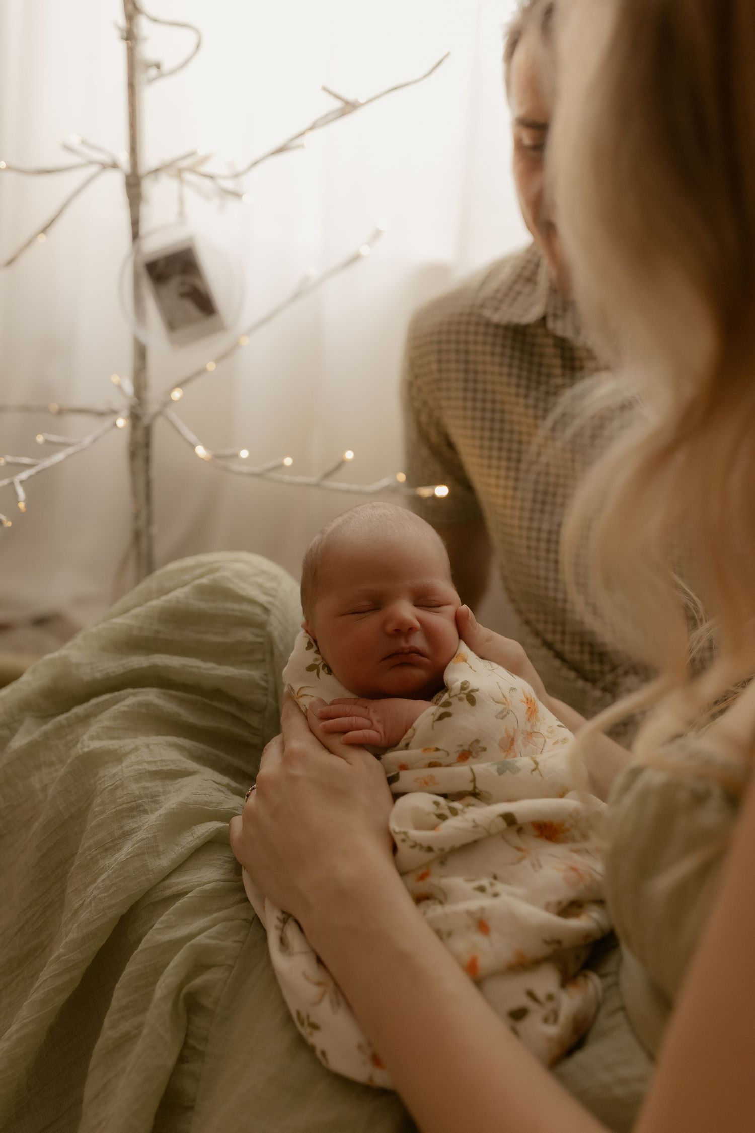 Gentle hands holding sleeping newborn under decorative branches and soft lighting.
