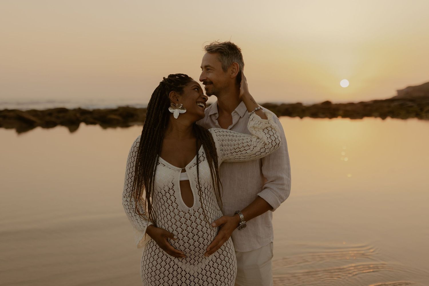A couple in white outfits embraces during sunset by a tranquil lake, creating a romantic silhouette against the golden sky.