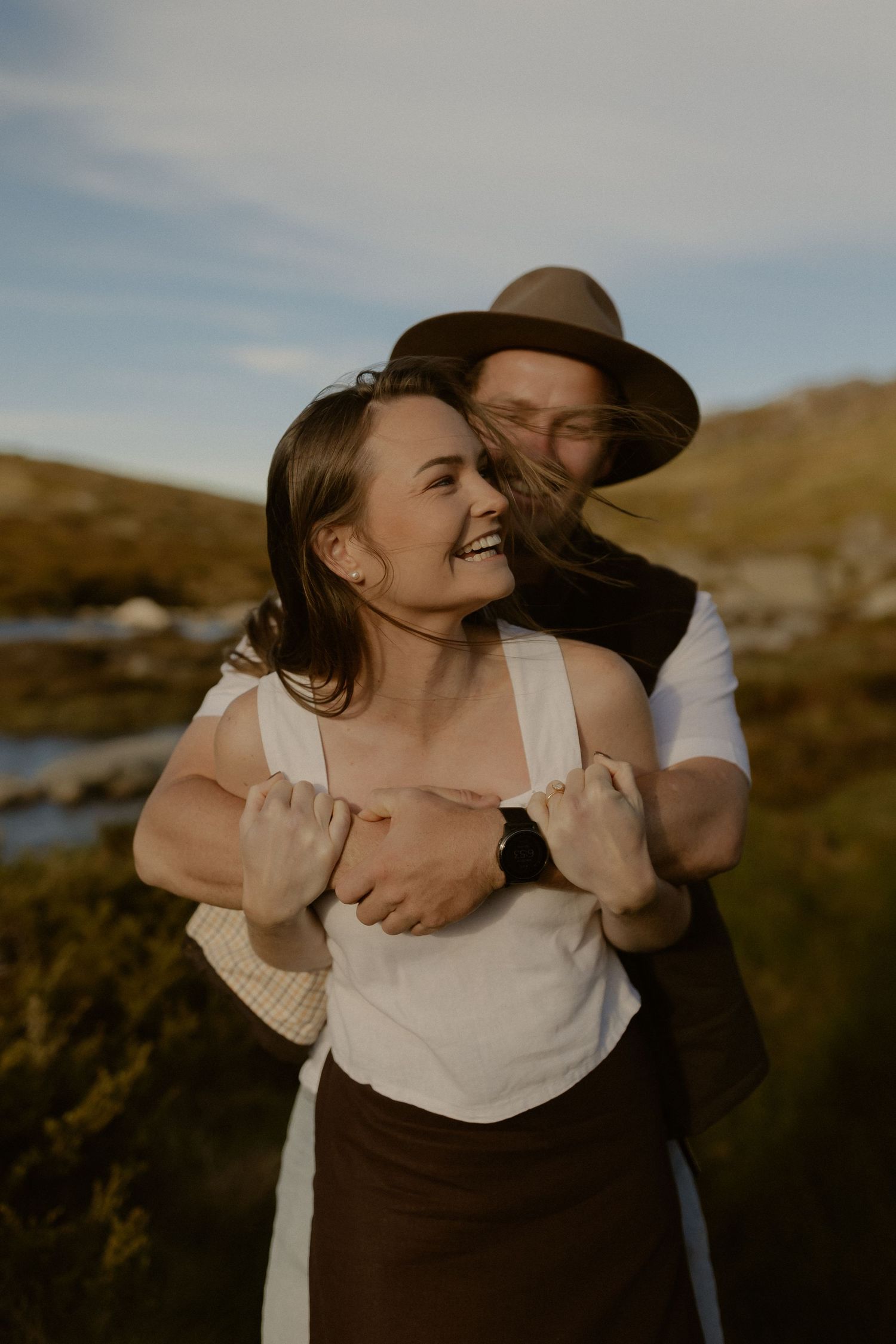 A couple embraces joyfully in a candid moment against a natural outdoor backdrop during golden hour.