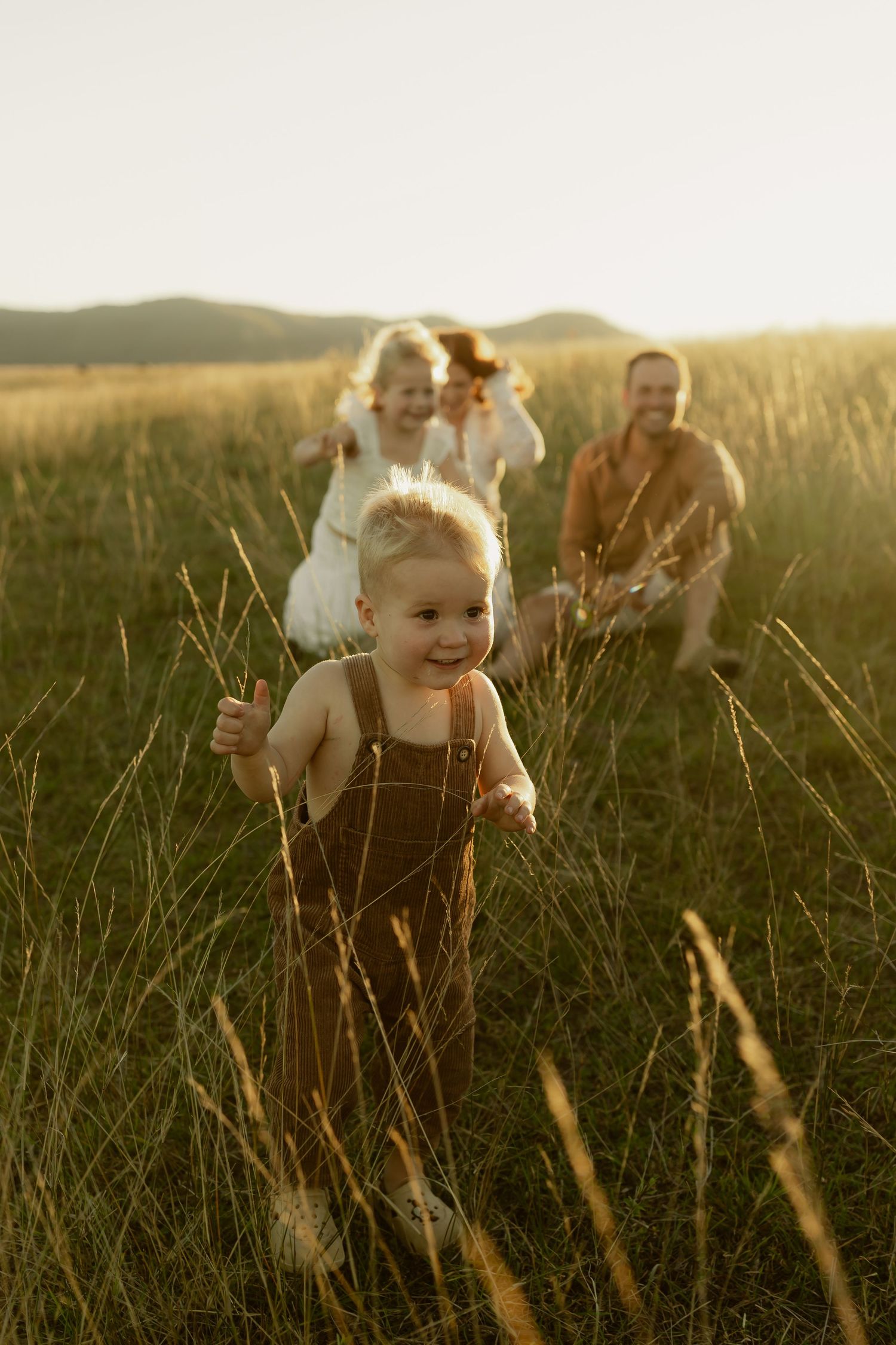 A happy toddler in brown overalls runs through tall grass as family plays in a golden field.