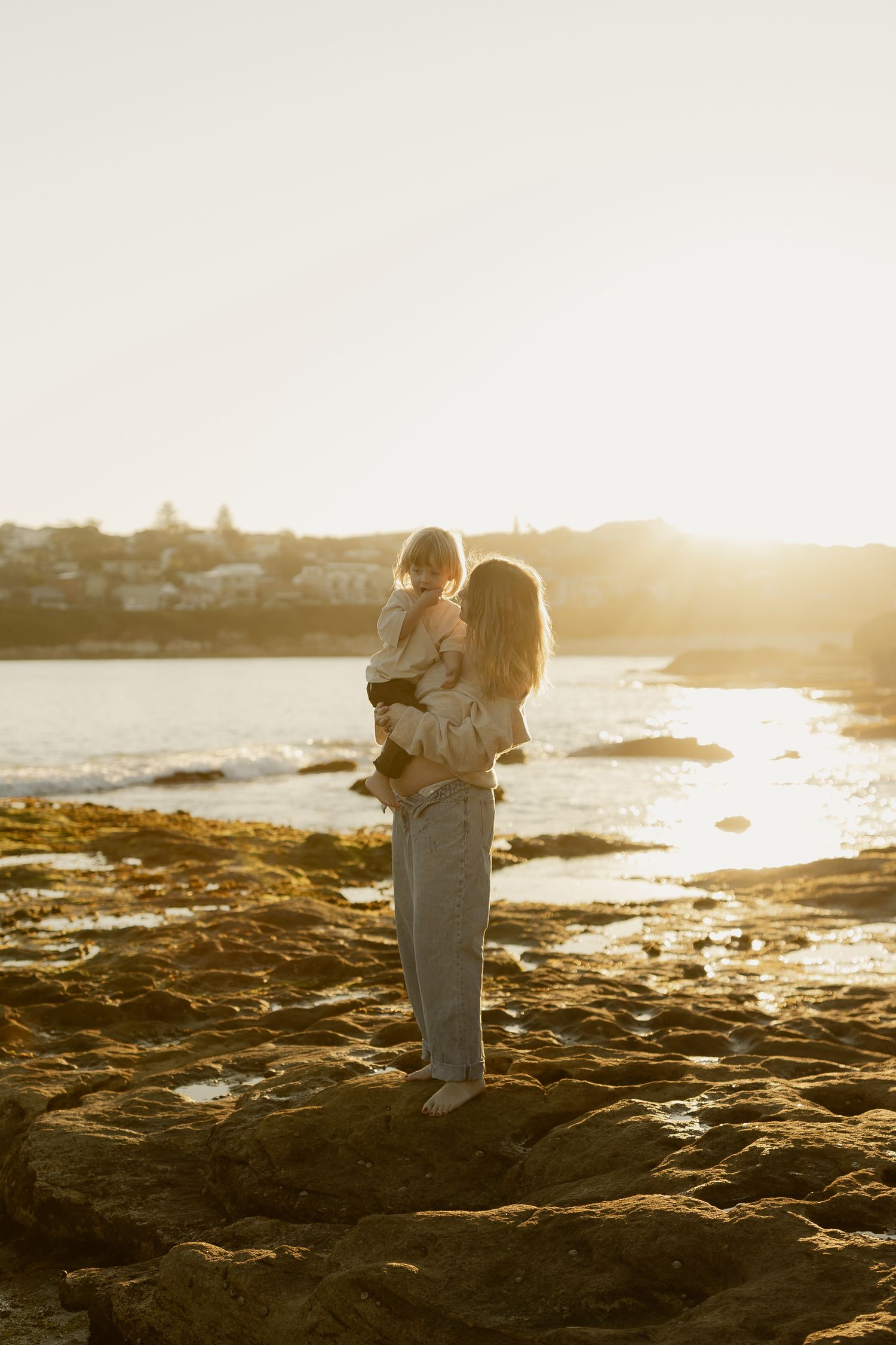 A mother holds her young child while standing on rocky shoreline at golden sunset with ocean and coastal town in background.