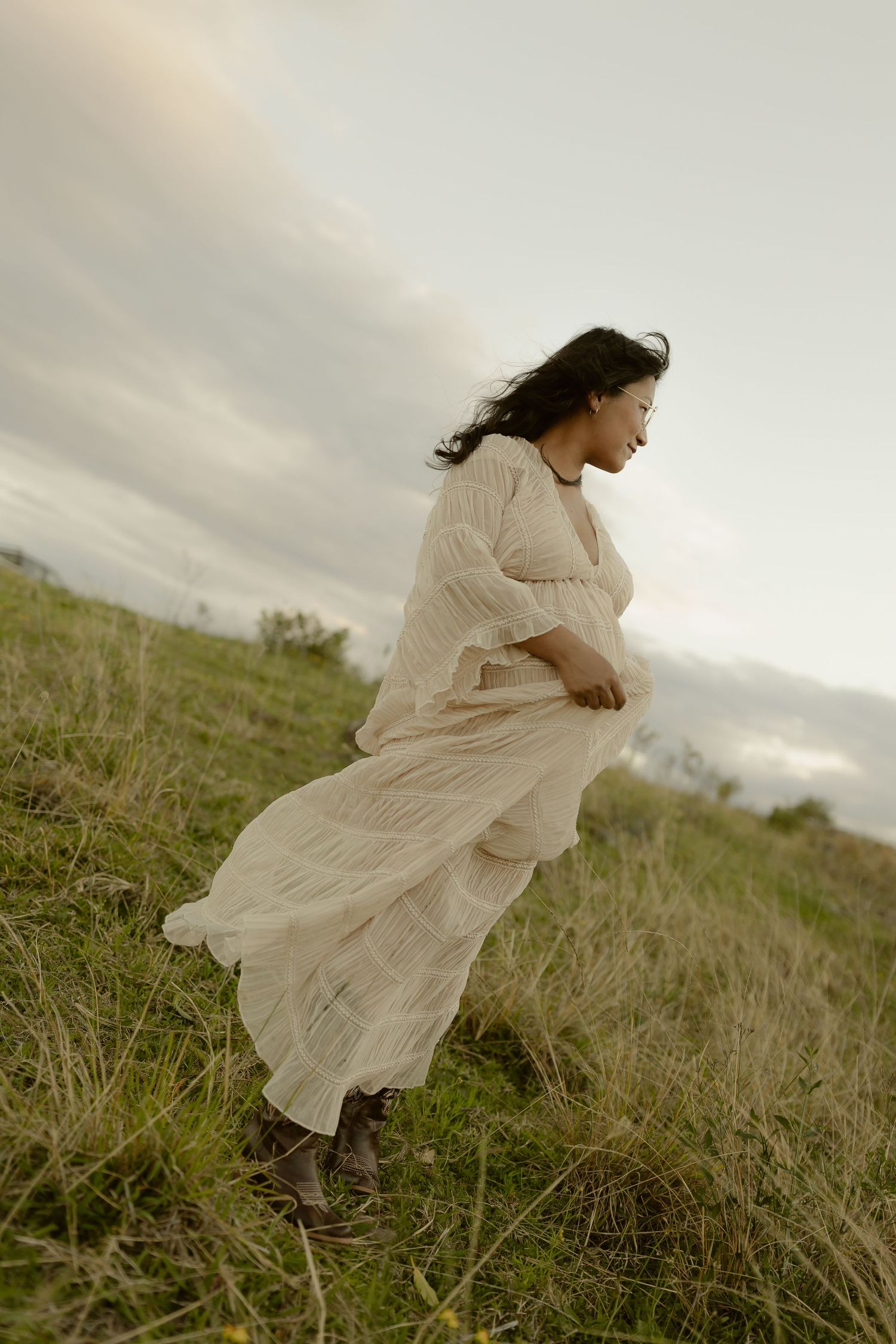 A woman in a flowing white dress walks through a grassy hillside, her hair and skirt blowing in the wind.