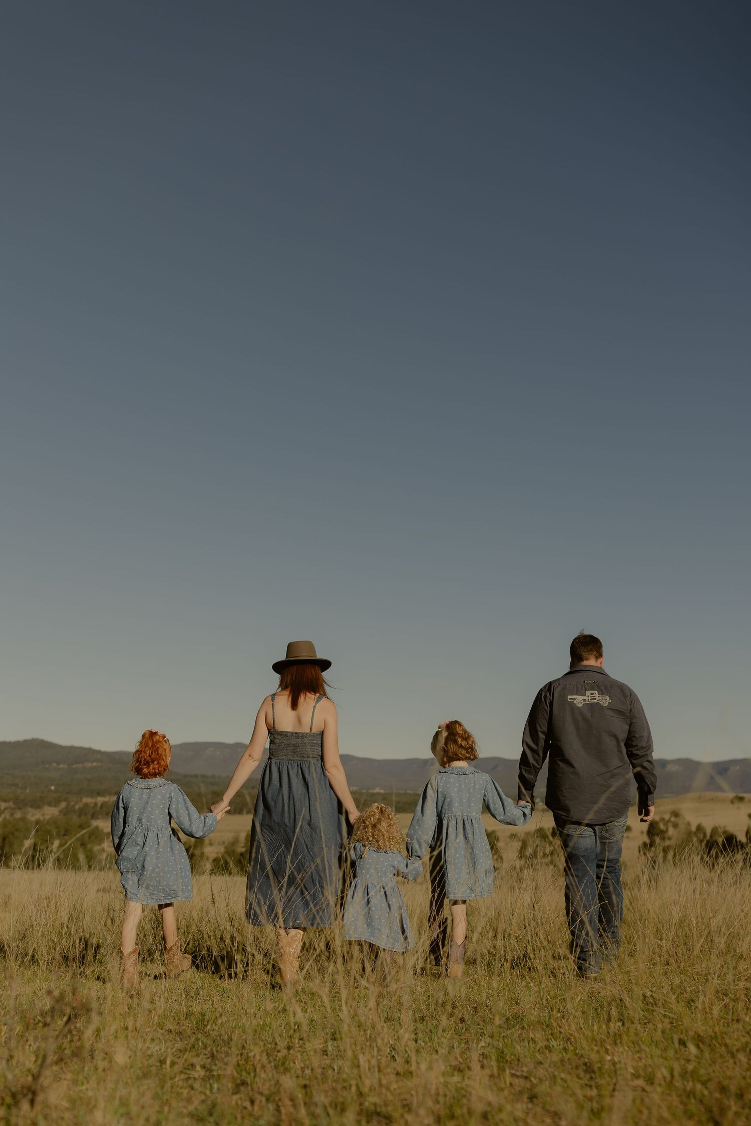 Family of five walking hand-in-hand through grassy field at sunset with mountain views in background.