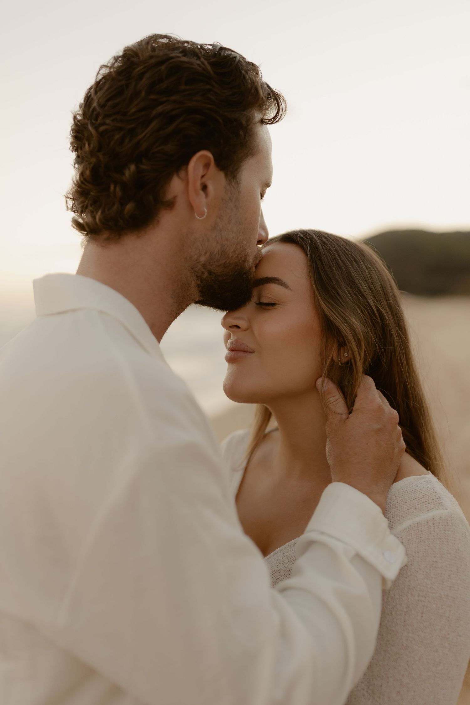 Romantic couple in white clothing embracing on beach during golden hour sunset.