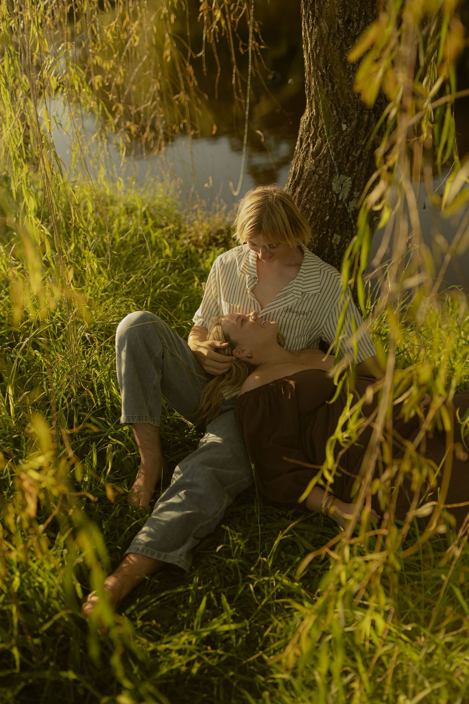 Person reading under willow tree branches in warm evening sunlight by water.