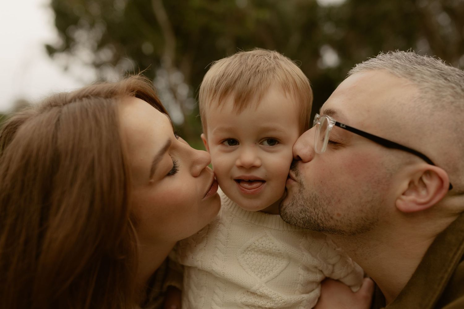 Parents kissing toddler on both cheeks in a joyful outdoor family portrait.