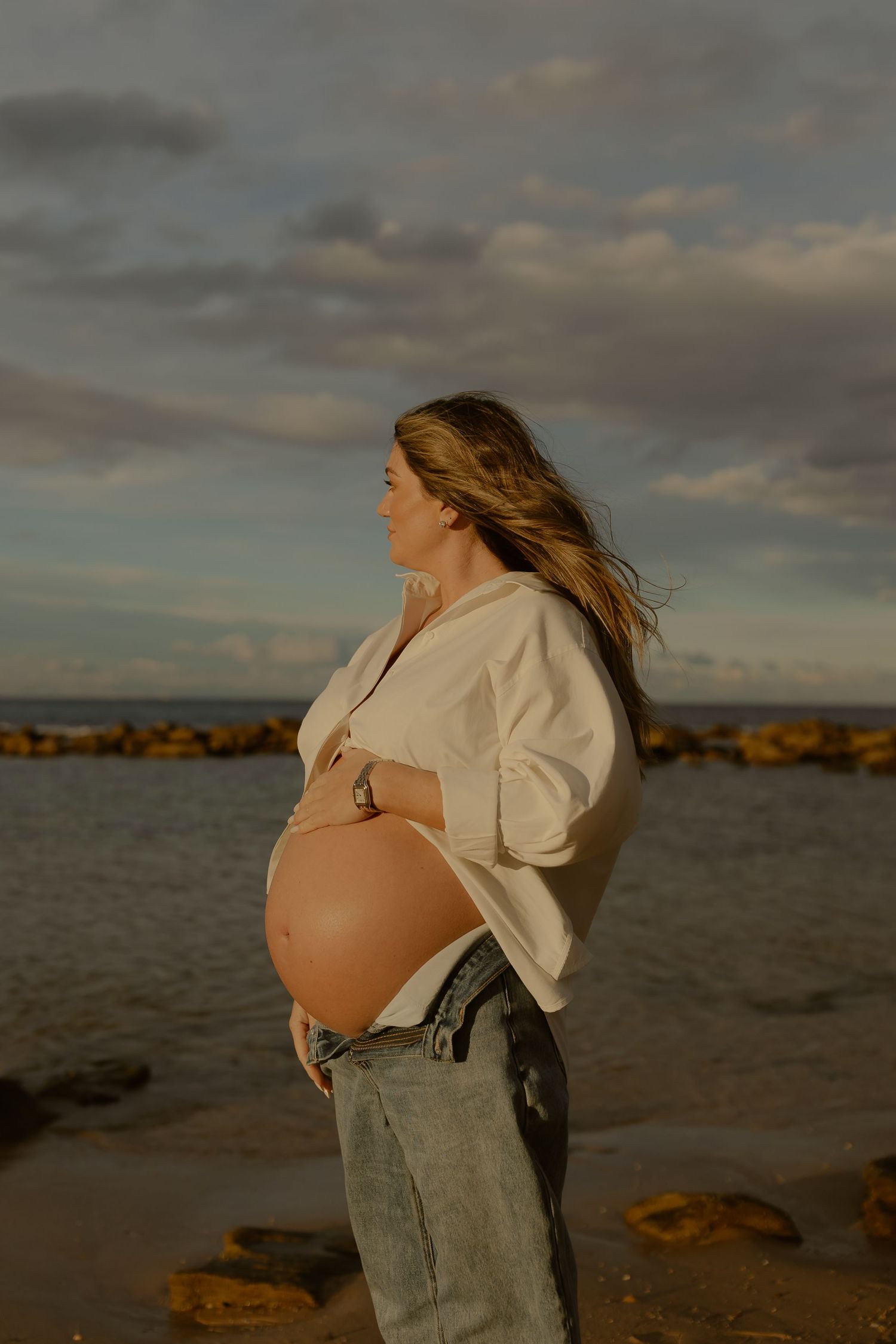 Pregnant woman in open white shirt cradles bare baby bump on a windy beach at golden hour.