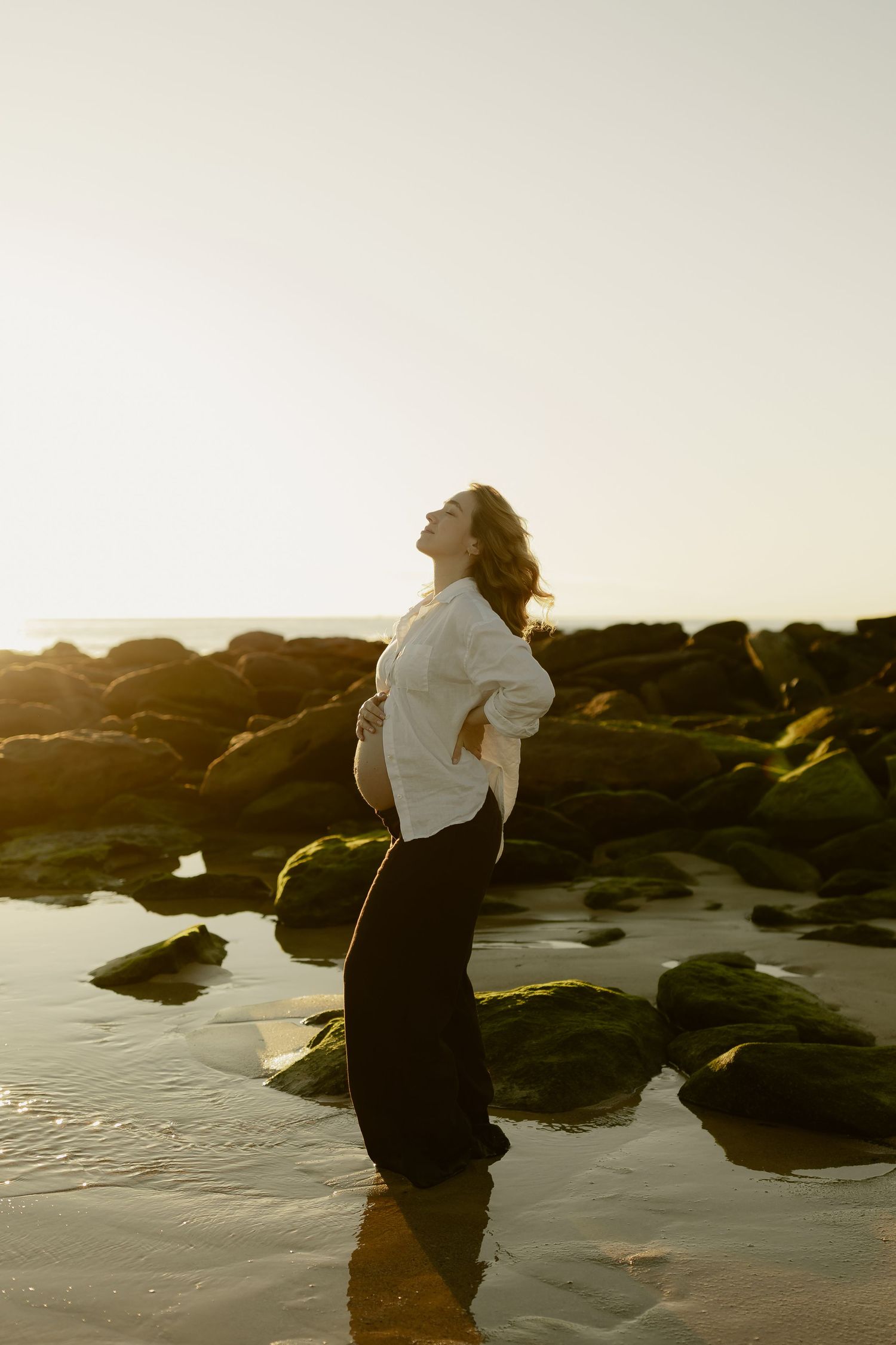 Pregnant woman in white shirt and black pants standing on rocky beach at sunset, cradling her baby bump.