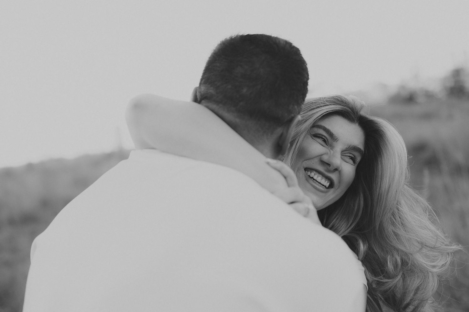 Black and white photo of a smiling woman embracing a man outdoors in a romantic couple portrait.