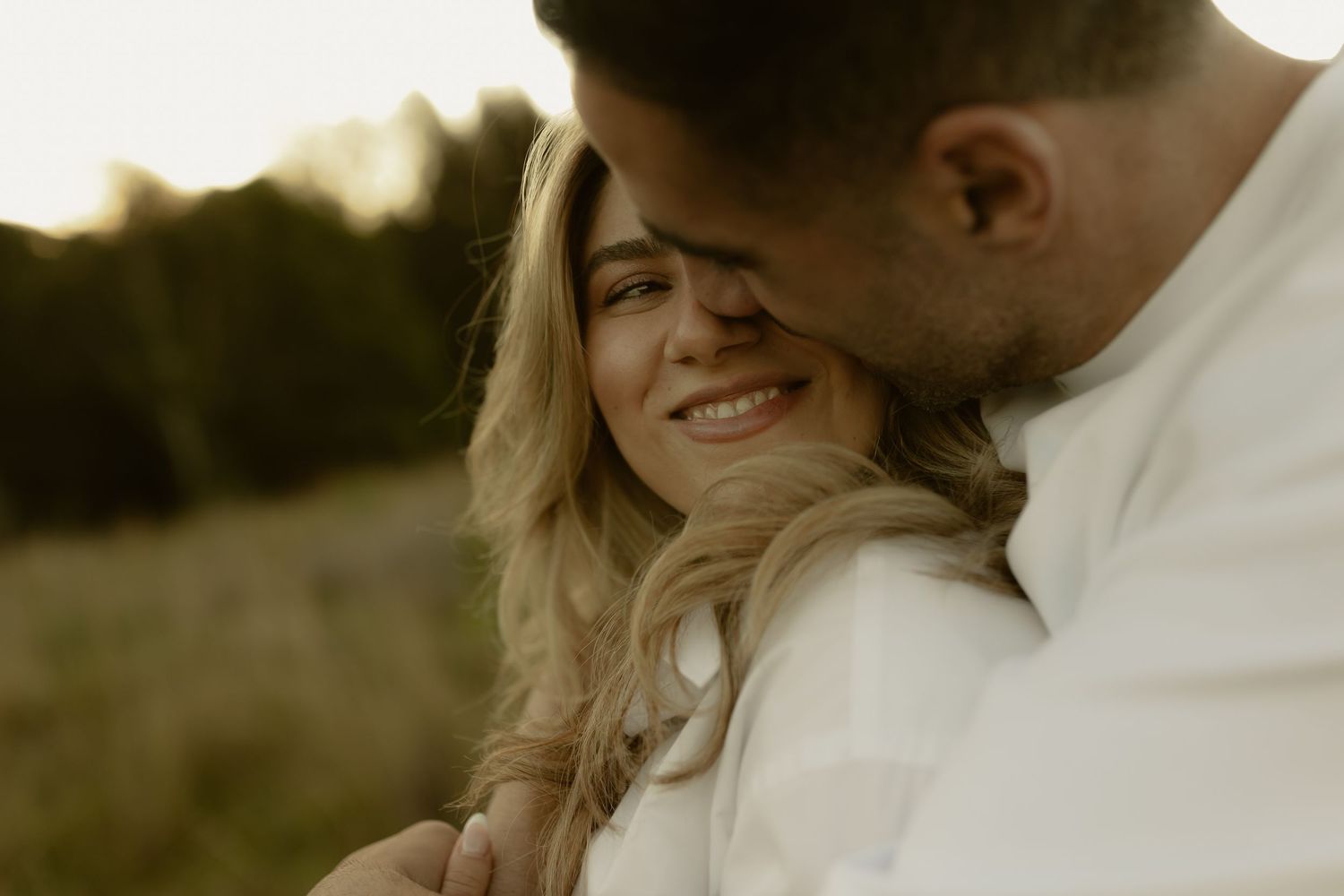 A couple shares a tender romantic moment outdoors at golden hour, woman smiling warmly as man leans close.