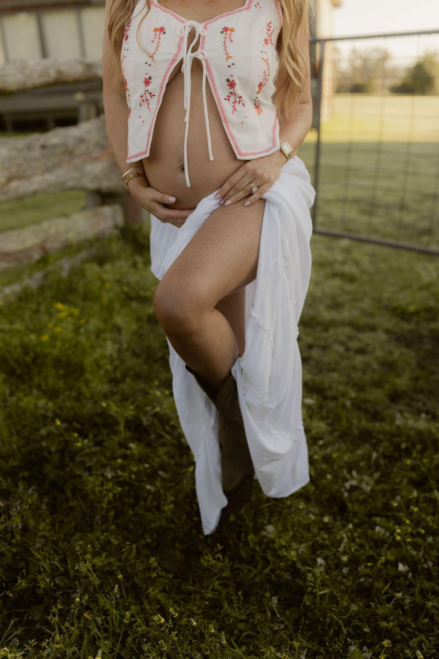 Pregnant woman in floral crop top and white skirt poses outdoors on green grass.
