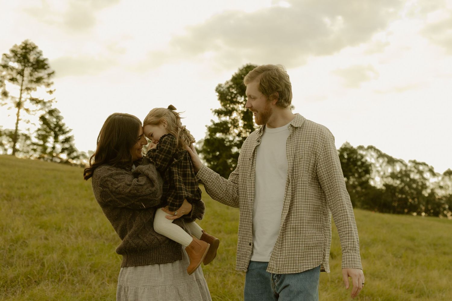 A young family with a toddler shares a tender moment in a lush green field at golden hour.