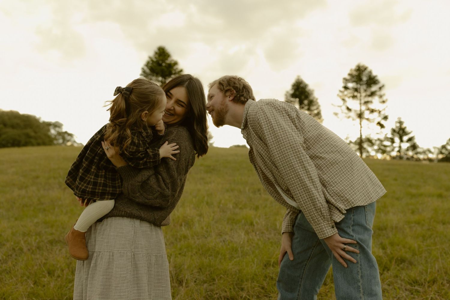 Family of three sharing a tender moment in a green field, mother holding daughter while father leans in for a kiss.