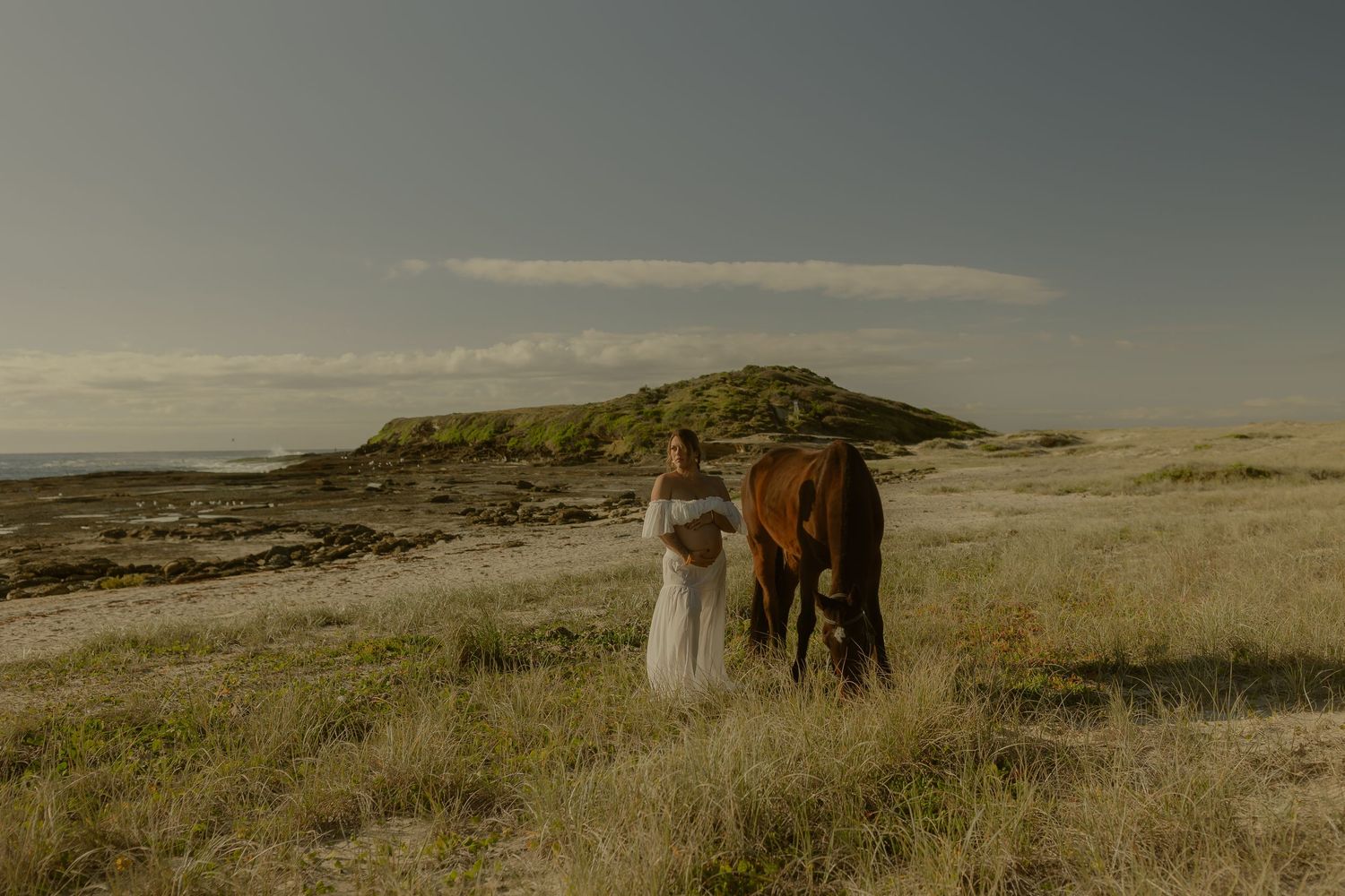 Bride in white dress standing beside a brown horse in a coastal landscape with sandy dunes and ocean views.