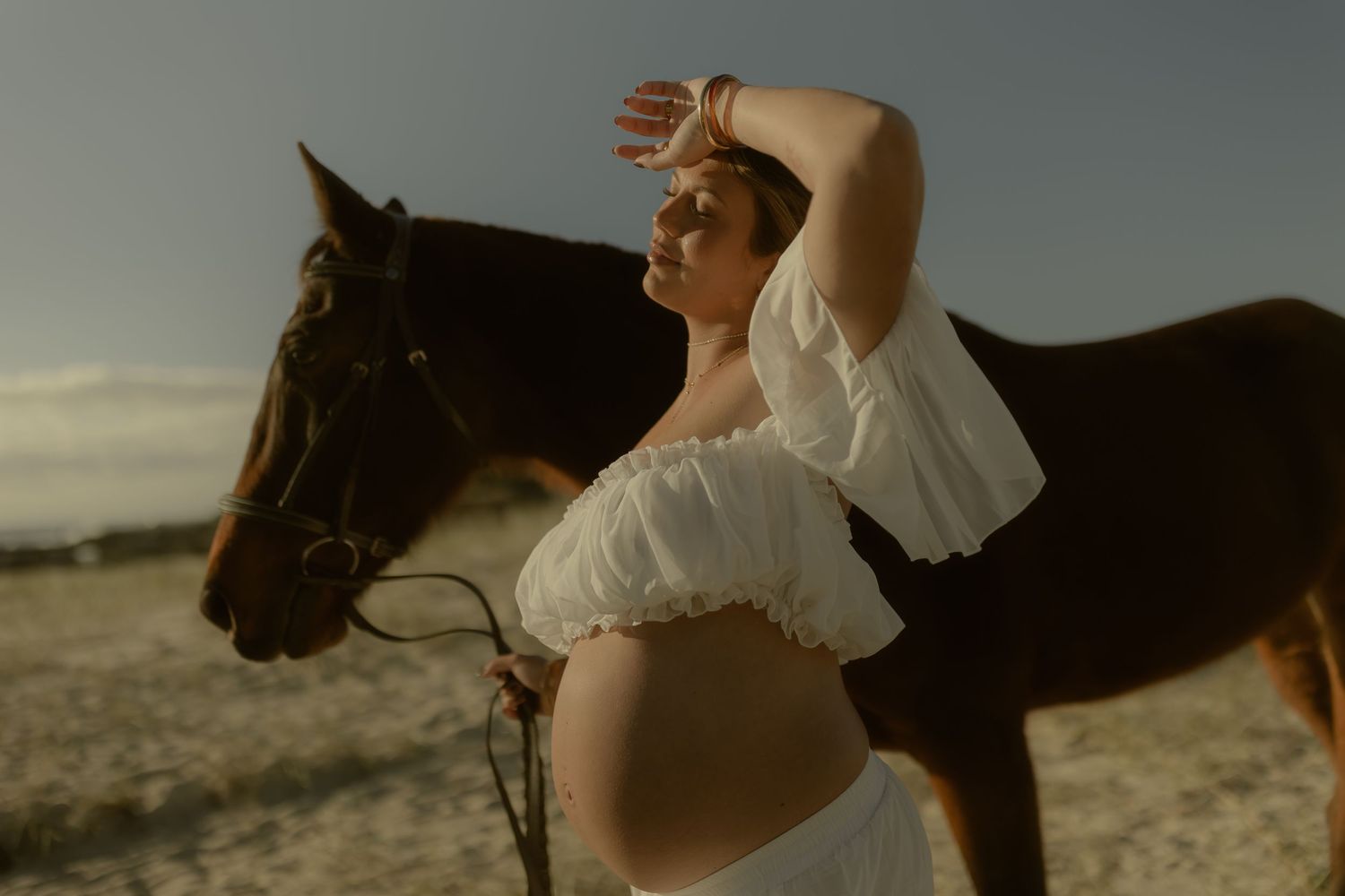 Pregnant woman in white crop top poses gracefully beside a brown horse on a sunlit sandy beach.