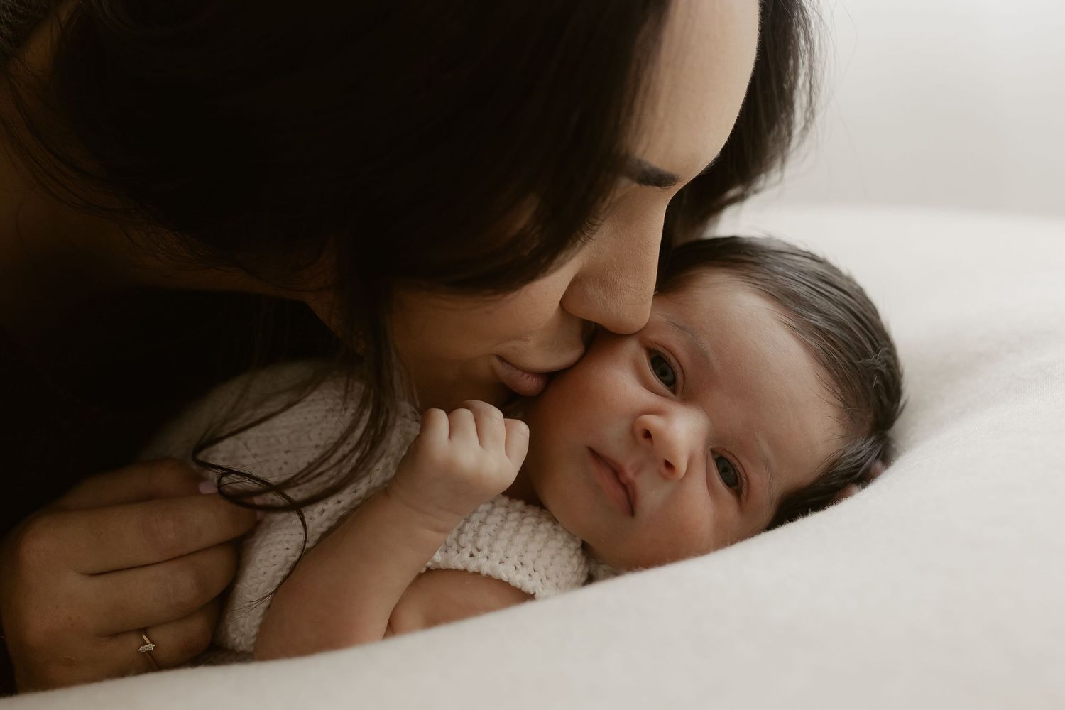 A mother tenderly kisses her newborn baby lying on a white blanket in a soft, intimate portrait.