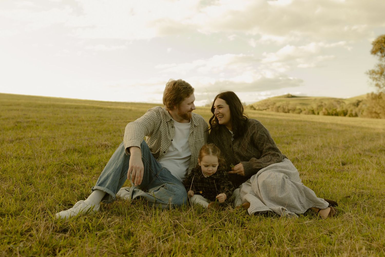 A happy family of three sits together on a lush green meadow, sharing a warm moment at golden hour.