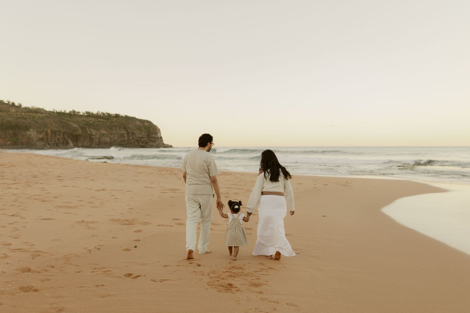 Family of three walking hand-in-hand on a sandy beach at sunset, wearing white outfits with ocean waves in the background.