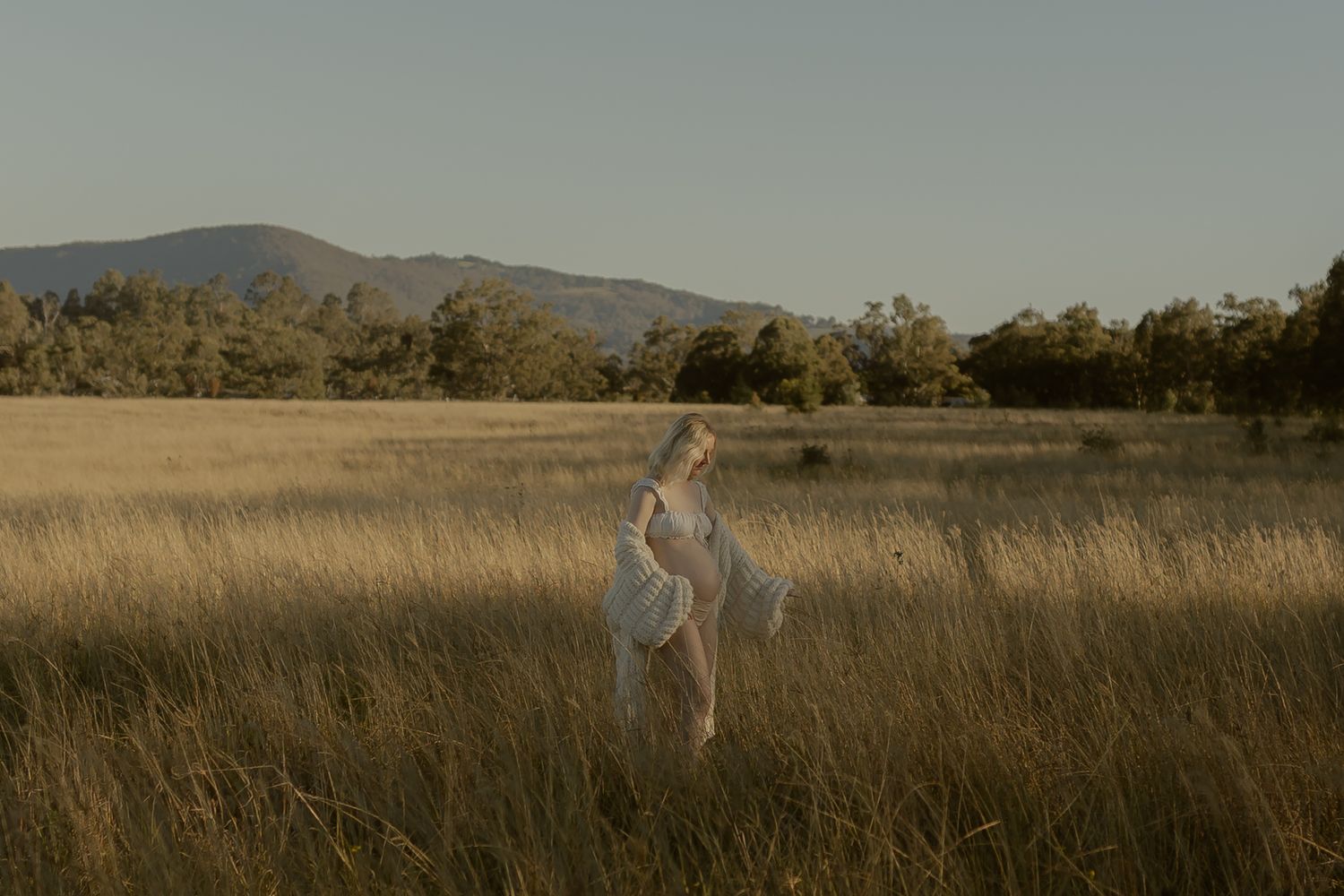 A woman in a flowing white dress stands gracefully in a golden field with mountains and trees in the background.