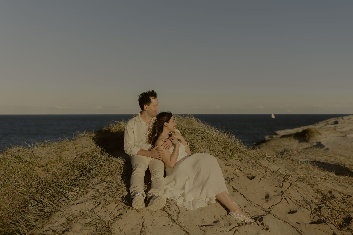 Couple sitting on sandy dunes by the ocean at golden hour, woman in white dress, man in casual attire.