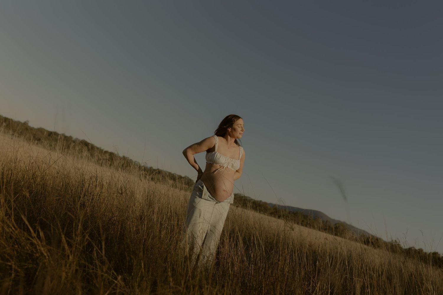 Pregnant woman in white dress standing in golden field at dusk with rolling hills in background.