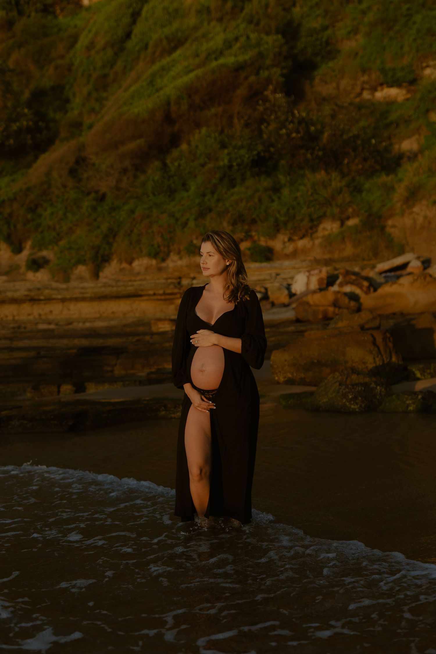Pregnant woman in black dress standing in ocean waves at golden hour with rocky cliffs behind her.