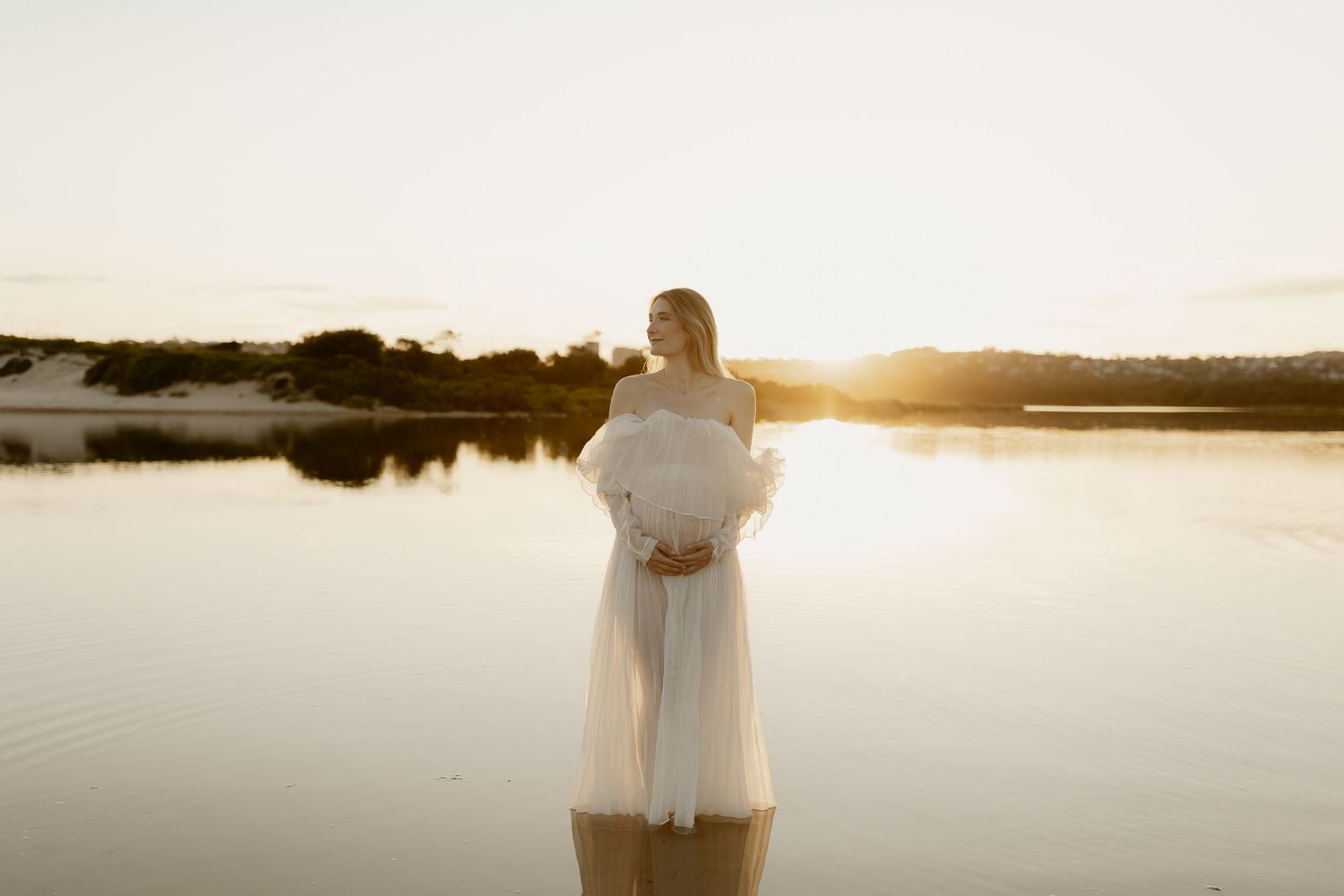 Silhouette of a person in white dress standing by tranquil waters at dusk.