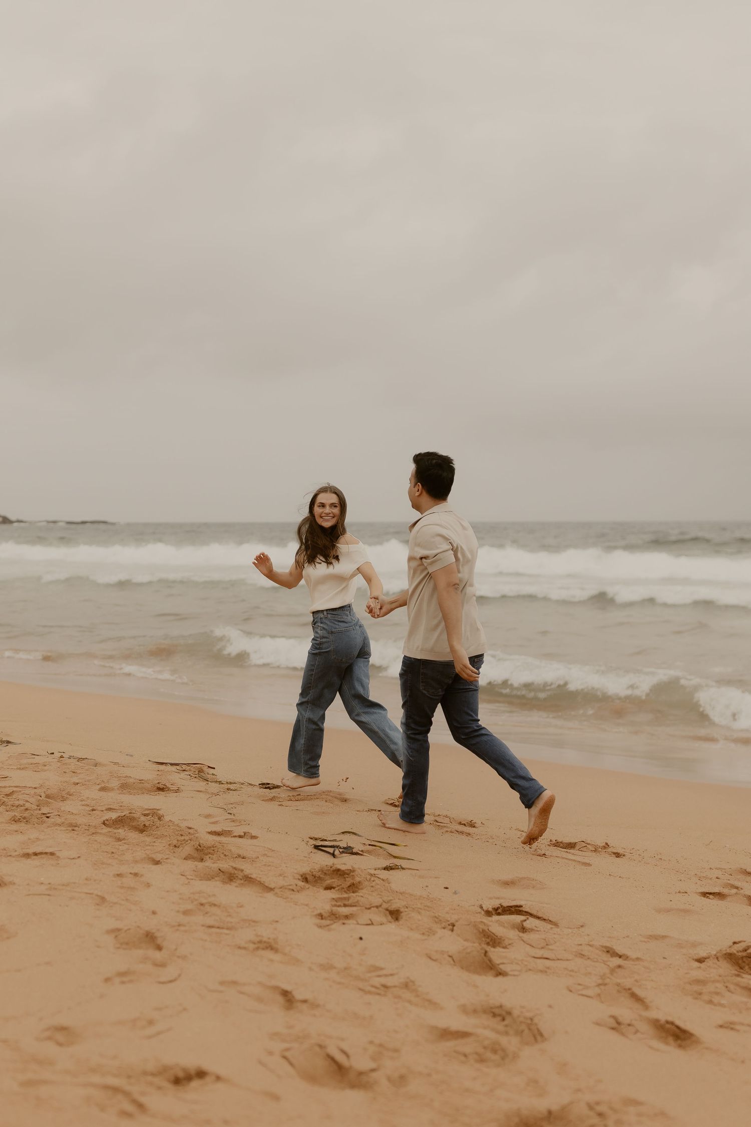 A playful couple runs and dances together on a misty beach.