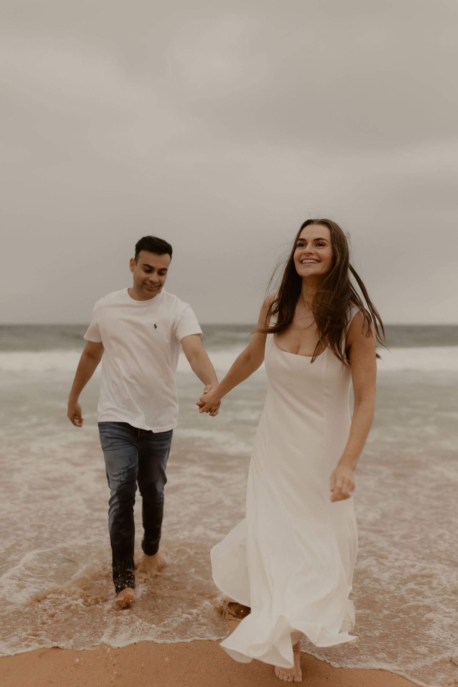 A couple walking hand in hand through gentle waves on an overcast beach.