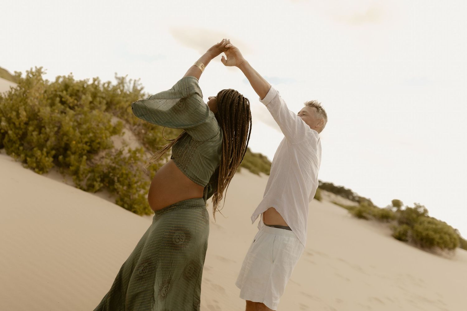 Two people walk through a sandy desert landscape during golden hour with sunlight creating long shadows.