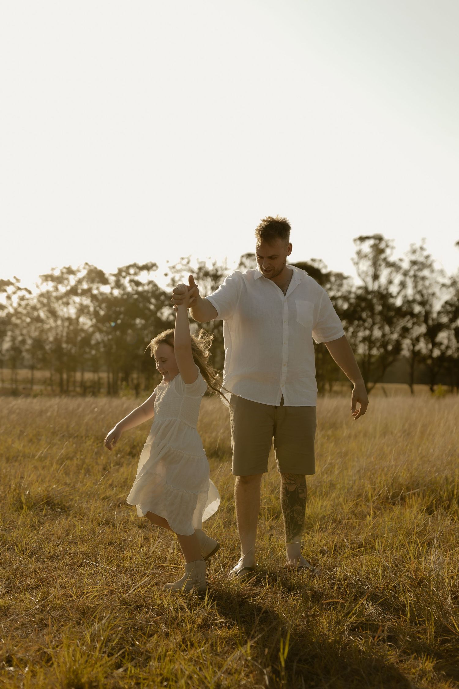 A couple in white clothing runs playfully through a sunlit meadow at golden hour.