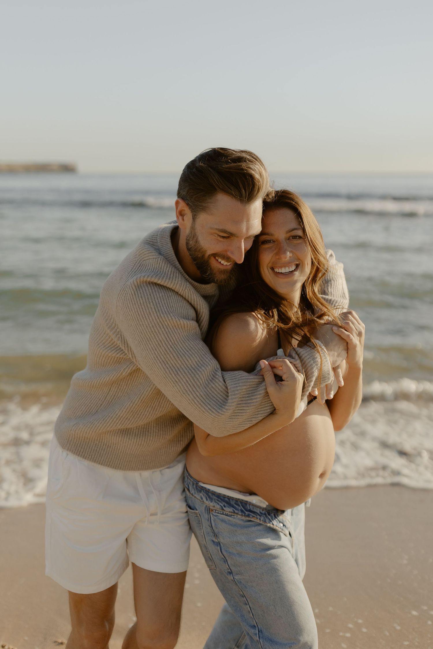 A couple shares joyful moments together on a sunny beach, wearing light colored clothing and embracing by the ocean waves.