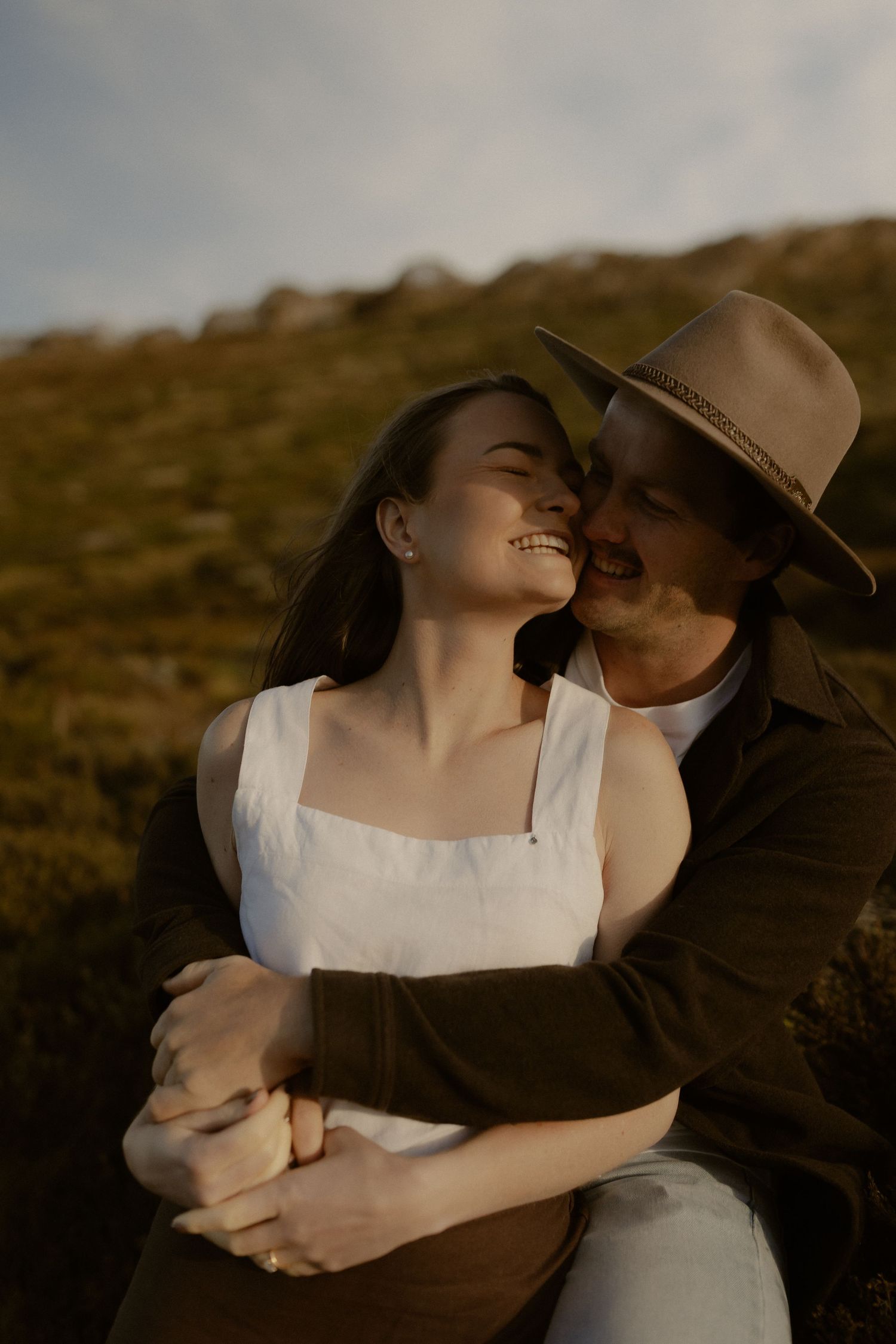 An intimate couple shares a tender moment on a hillside at sunset, with one wearing a brown hat and white dress.