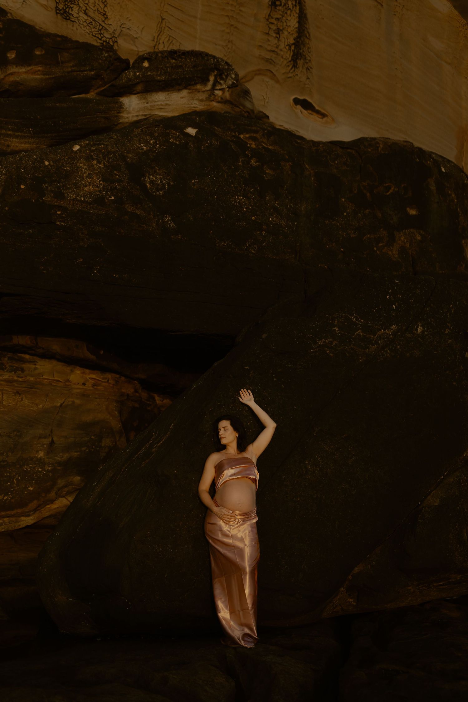 Figure in flowing silk dress poses dramatically against dark cave wall in moody artistic portrait.