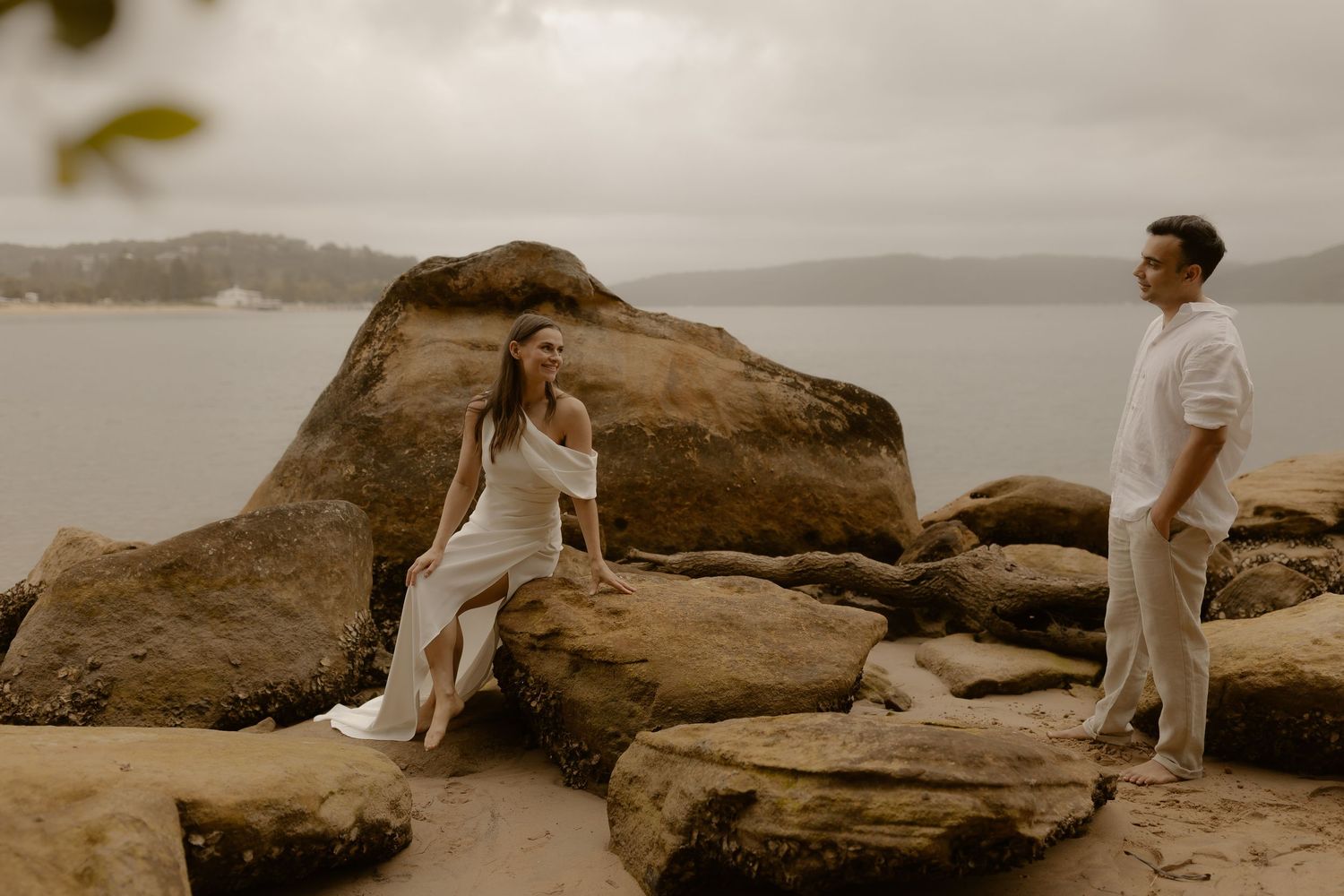 Two people in light clothing embrace while standing on large rocky outcrops by the ocean.