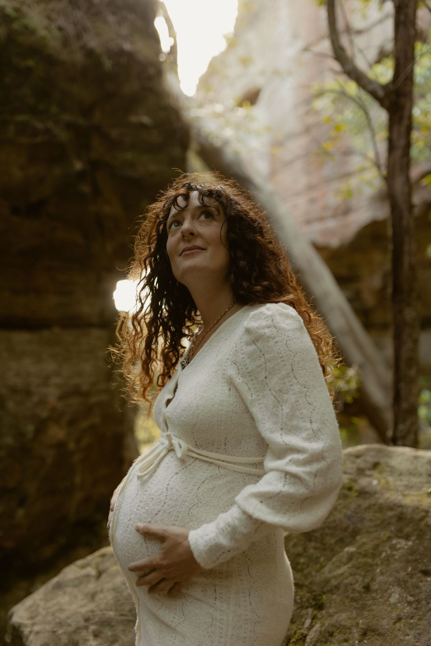 A dreamy maternity photoshoot in nature shows a pregnant woman in white lace dress posing against sunlit rock formations.