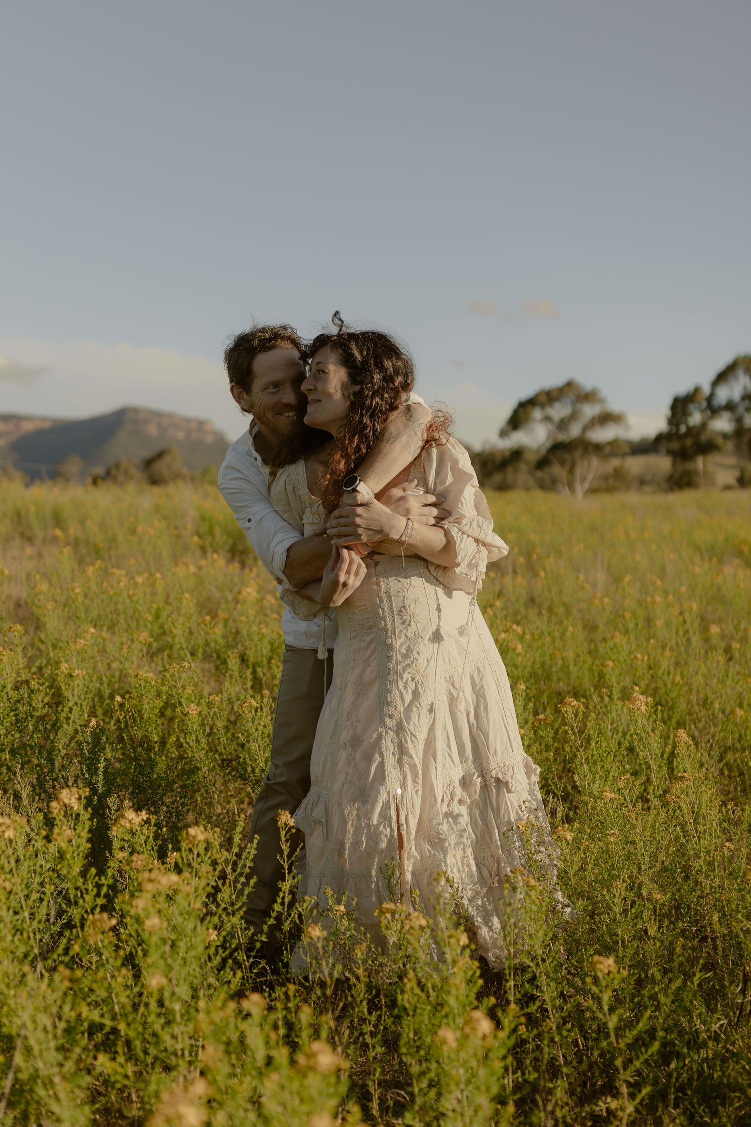 A romantic couple embraces in a sunlit meadow with mountains in the background during golden hour.