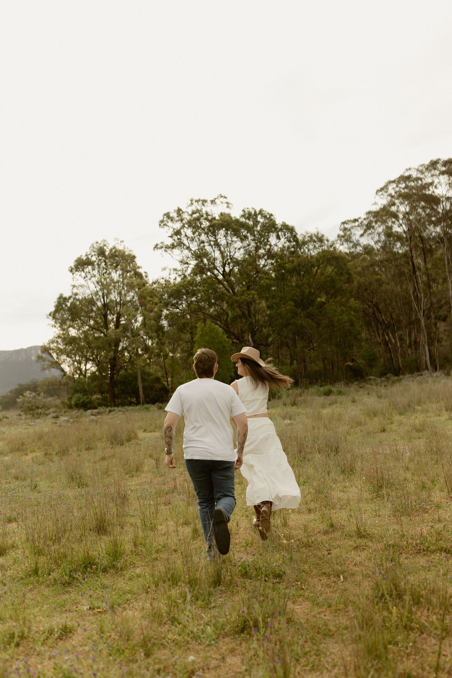 A couple walks hand in hand through a grassy field with eucalyptus trees in the distance.