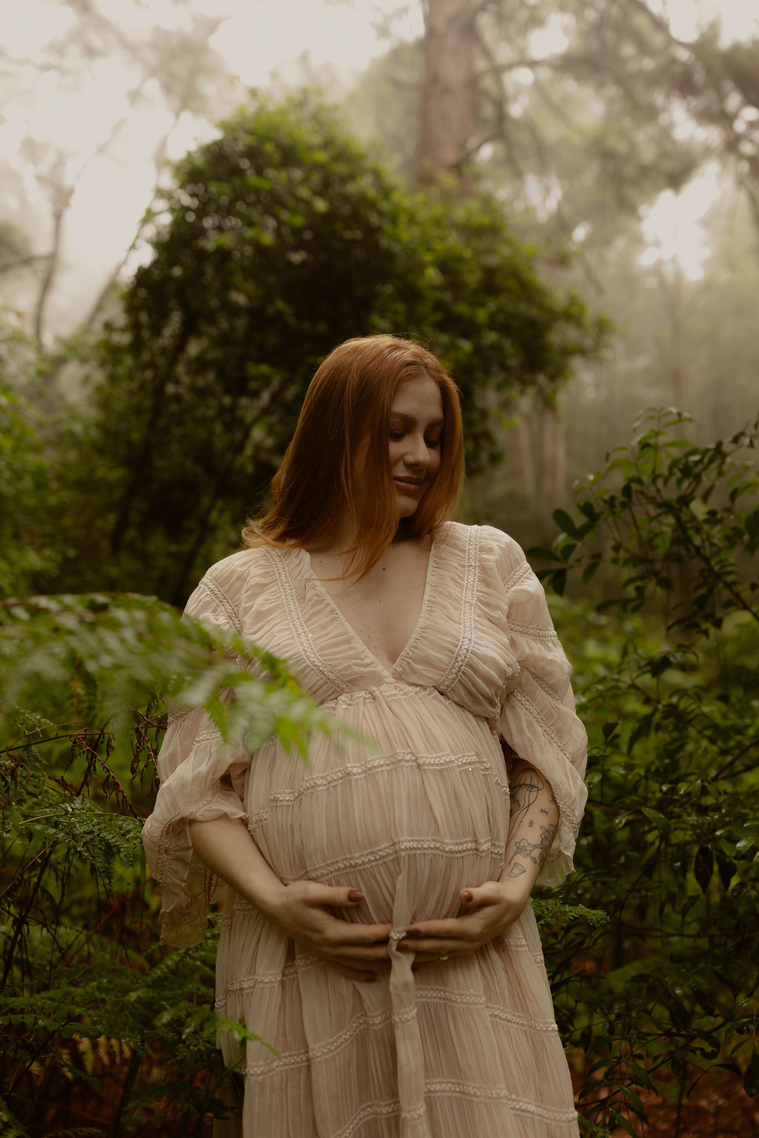 A romantic maternity photoshoot in a misty forest features a white lace dress and ethereal atmosphere.