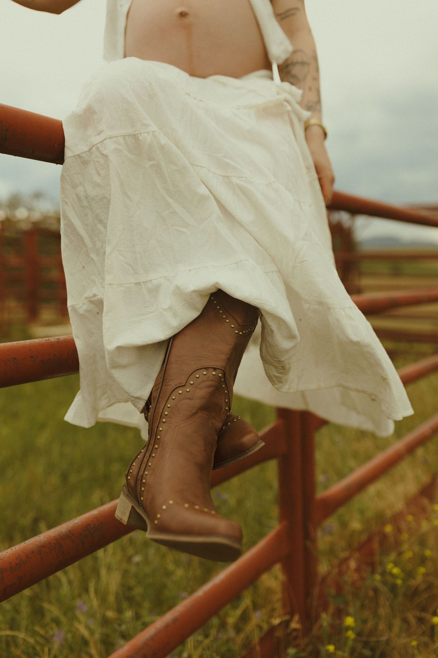 Brown leather boots peek out from under a flowing white dress while sitting on a rustic fence.