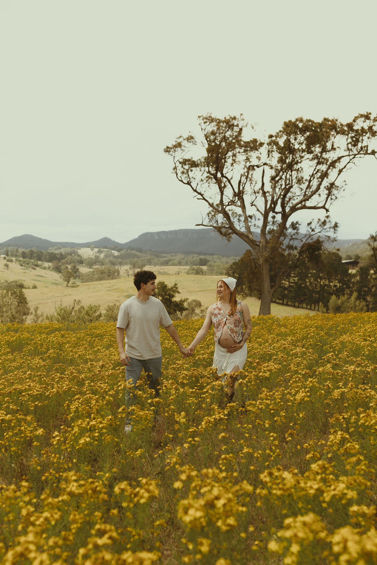 Couple walking hand in hand through a vast meadow of yellow flowers with mountains in background.