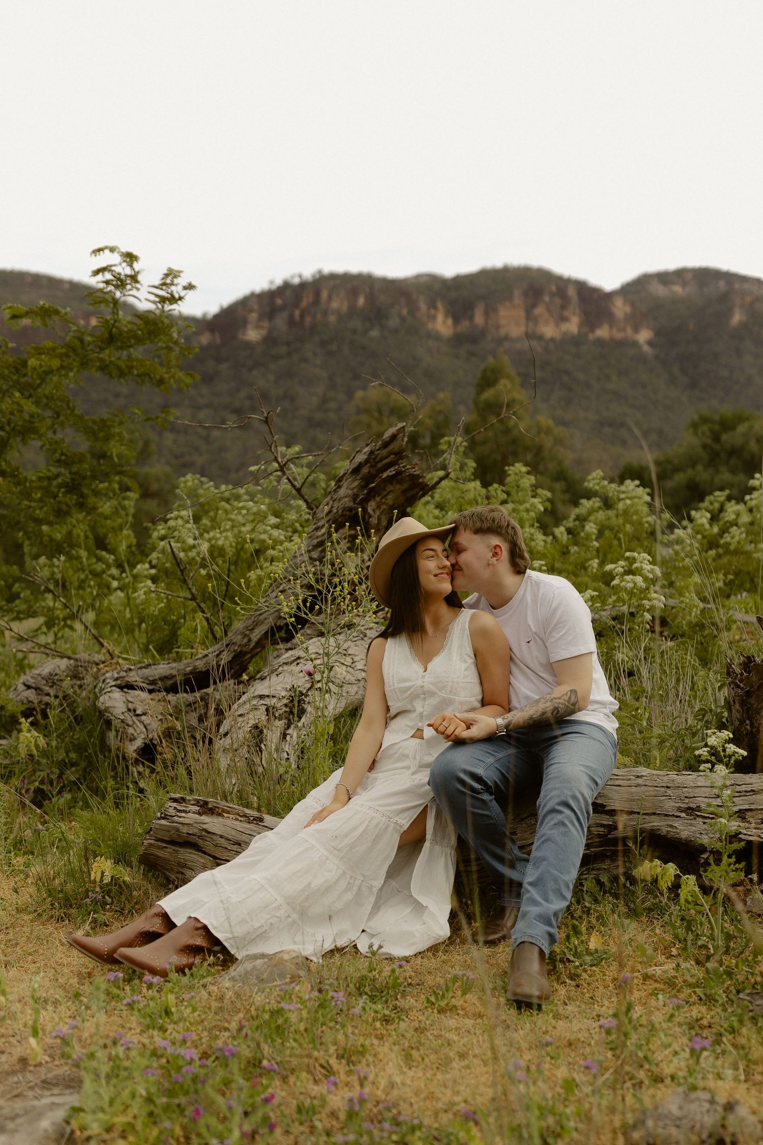 A couple shares a romantic moment while sitting on a log in a scenic mountain meadow filled with wildflowers at sunset.